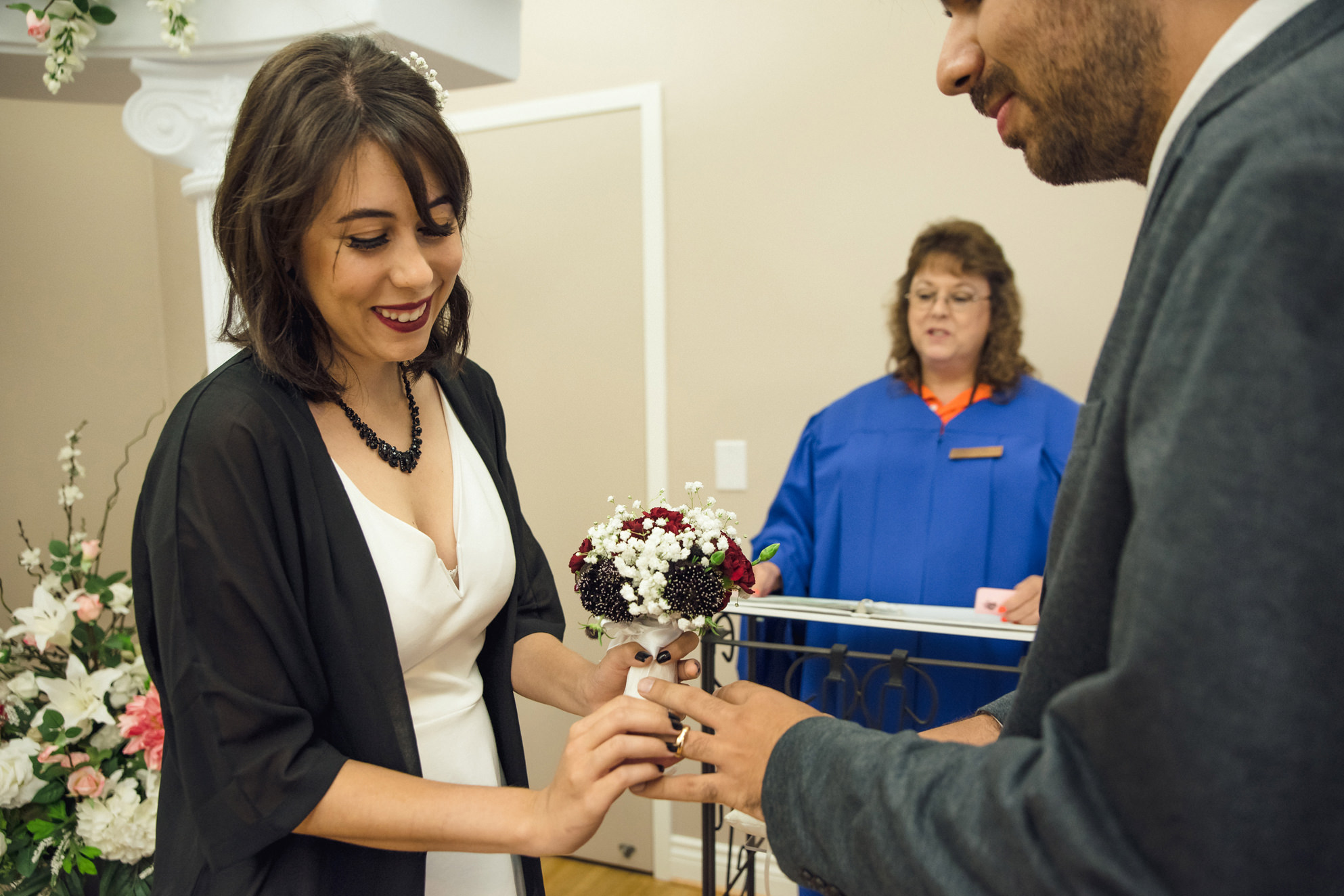 A bride smiling while putting a ring on her husband's finger at a ceremony at the San Jose Clerk Recording Office backyard