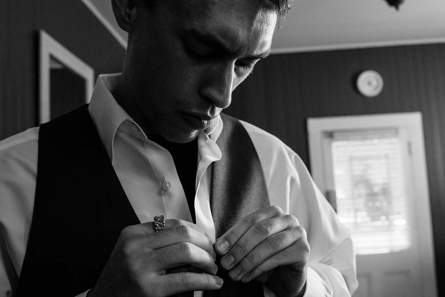 A Black and White image of a groom buttoning his wedding dress shirt