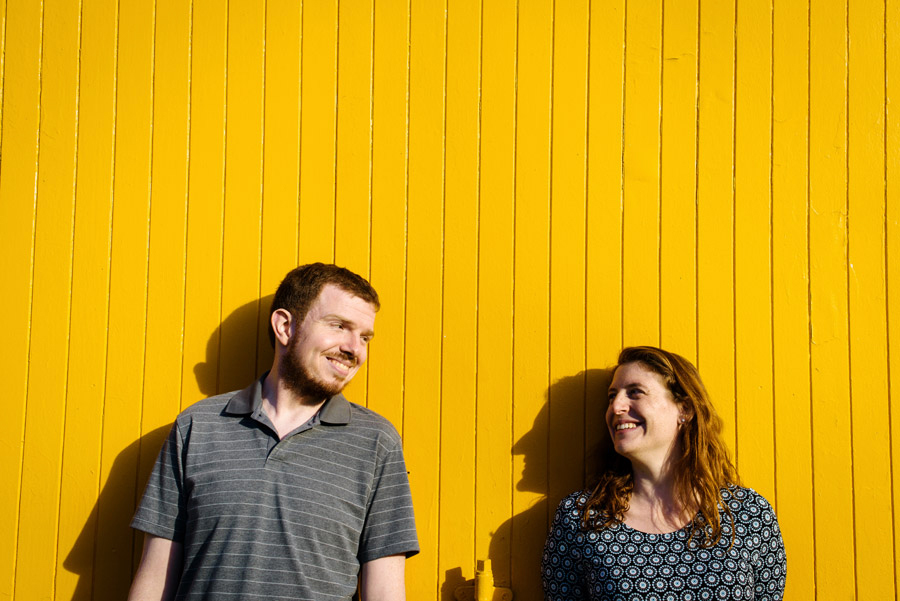 A happy couple stands on a yellow wooden wall looking at each other